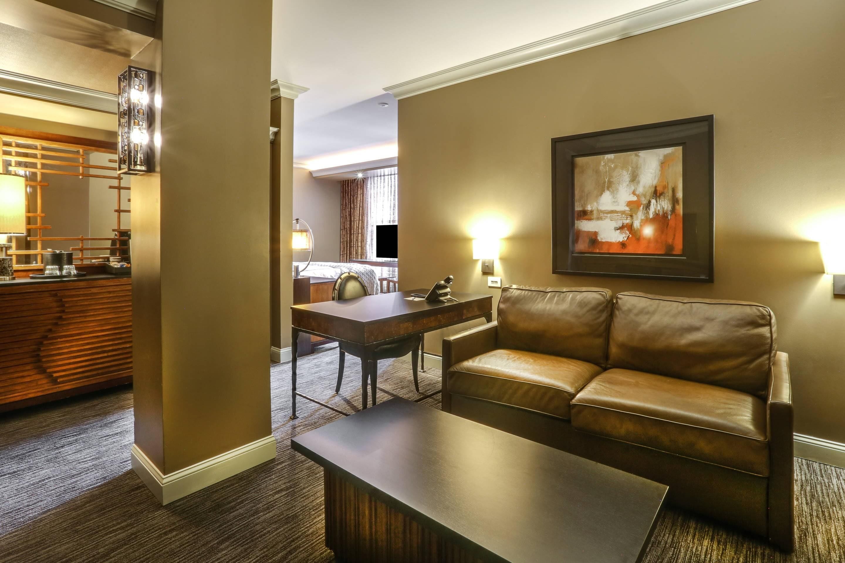 A hotel room with a leather sofa, a coffee table, a dining area with a table and chairs, and a painting on the wall.