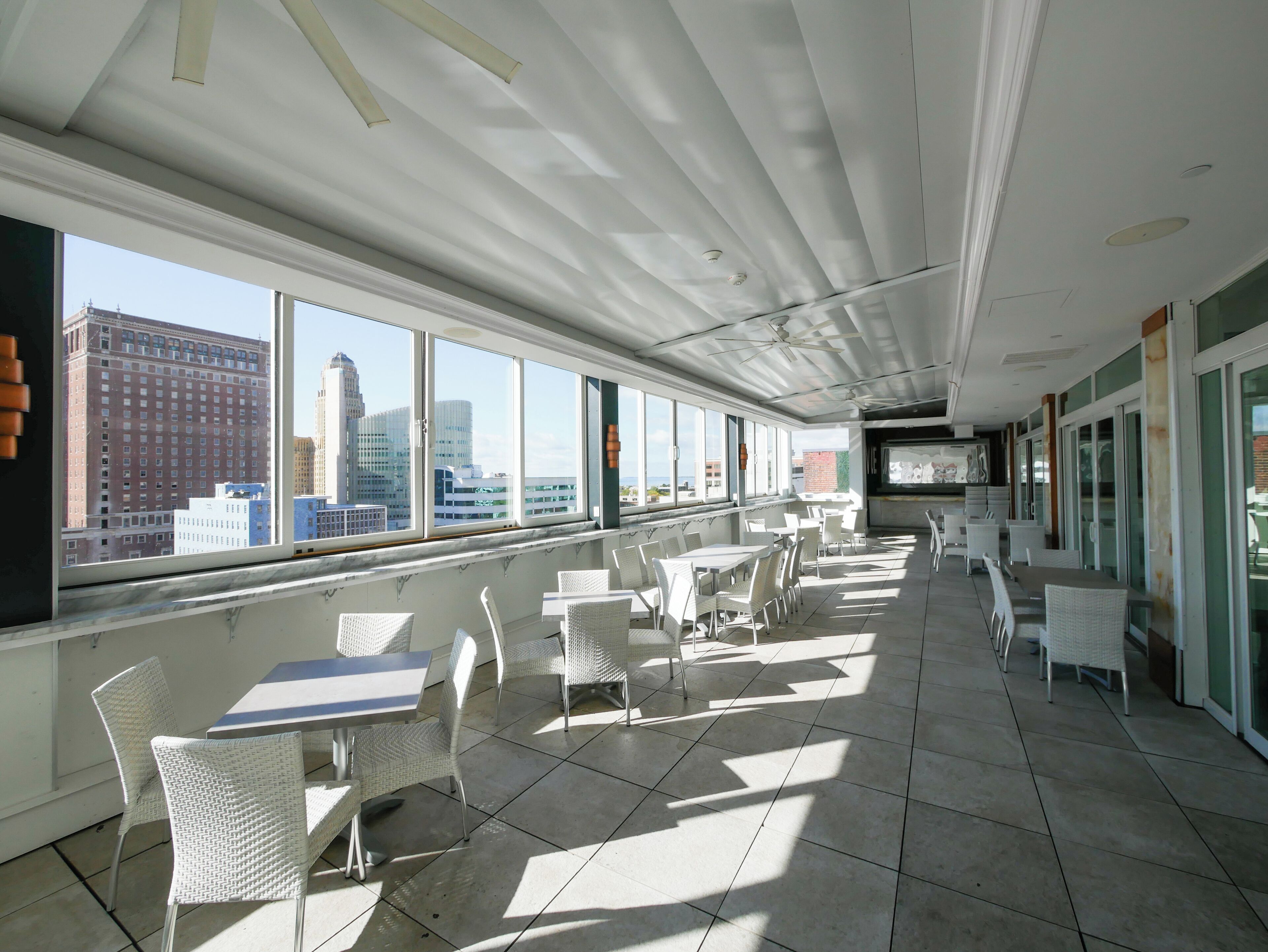 Un espace intérieur vaste et lumineux, avec de grandes fenêtres donnant sur la ville, des meubles en osier blancs et un sol carrelé.