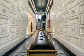 A hallway with a black leather bench, white decorative panels, a chandelier, and a staircase. - Curtiss Hotel, an Ascend Collection Hotel (Buffalo)