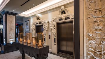Un lobby ornamentado con una larga mesa que exhibe velas y cristalería, un ascensor y detallados relieves en las paredes.