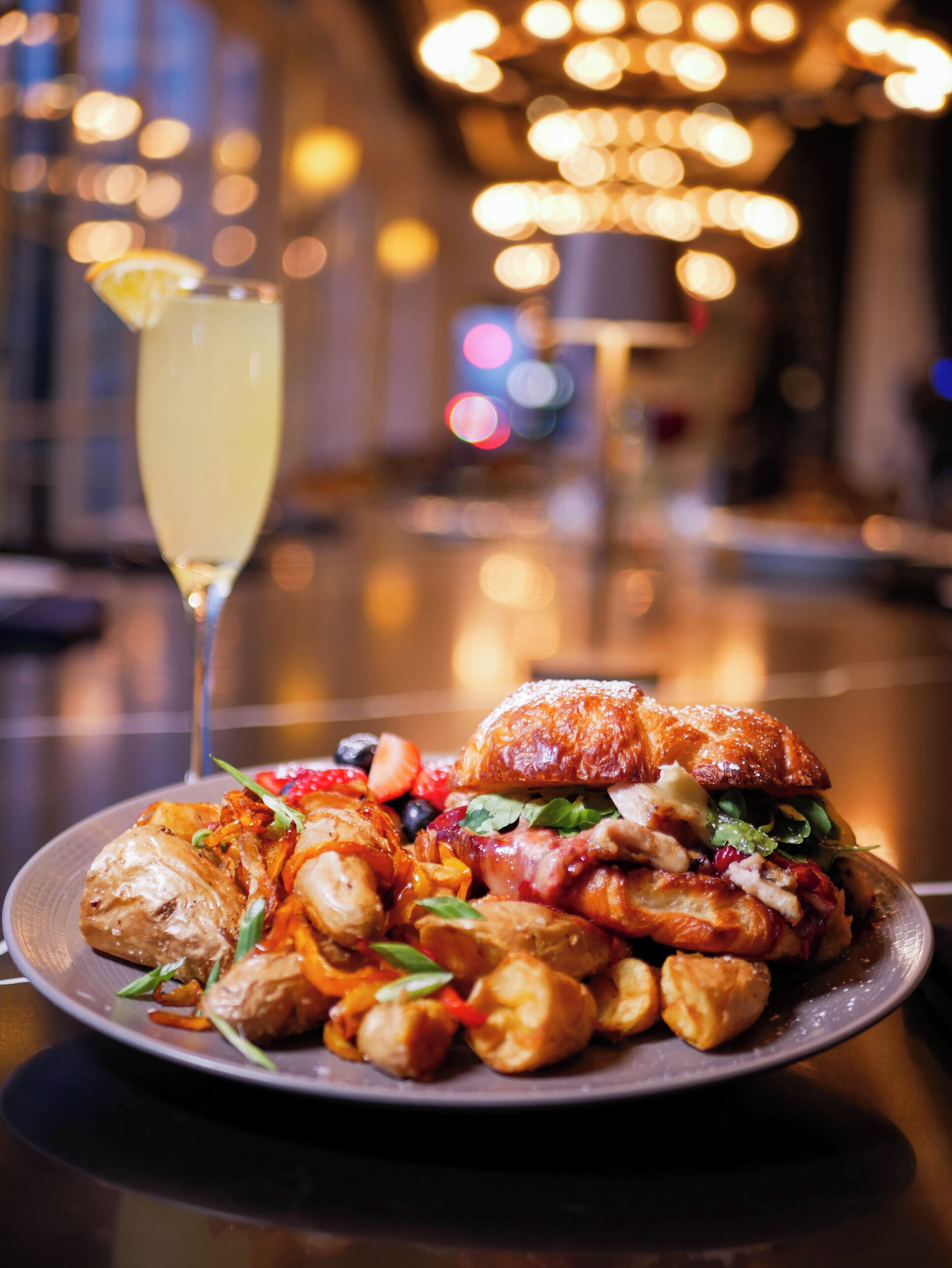 A plate of food with a sandwich and vegetables, a glass of drink, and a blurred background with lights.