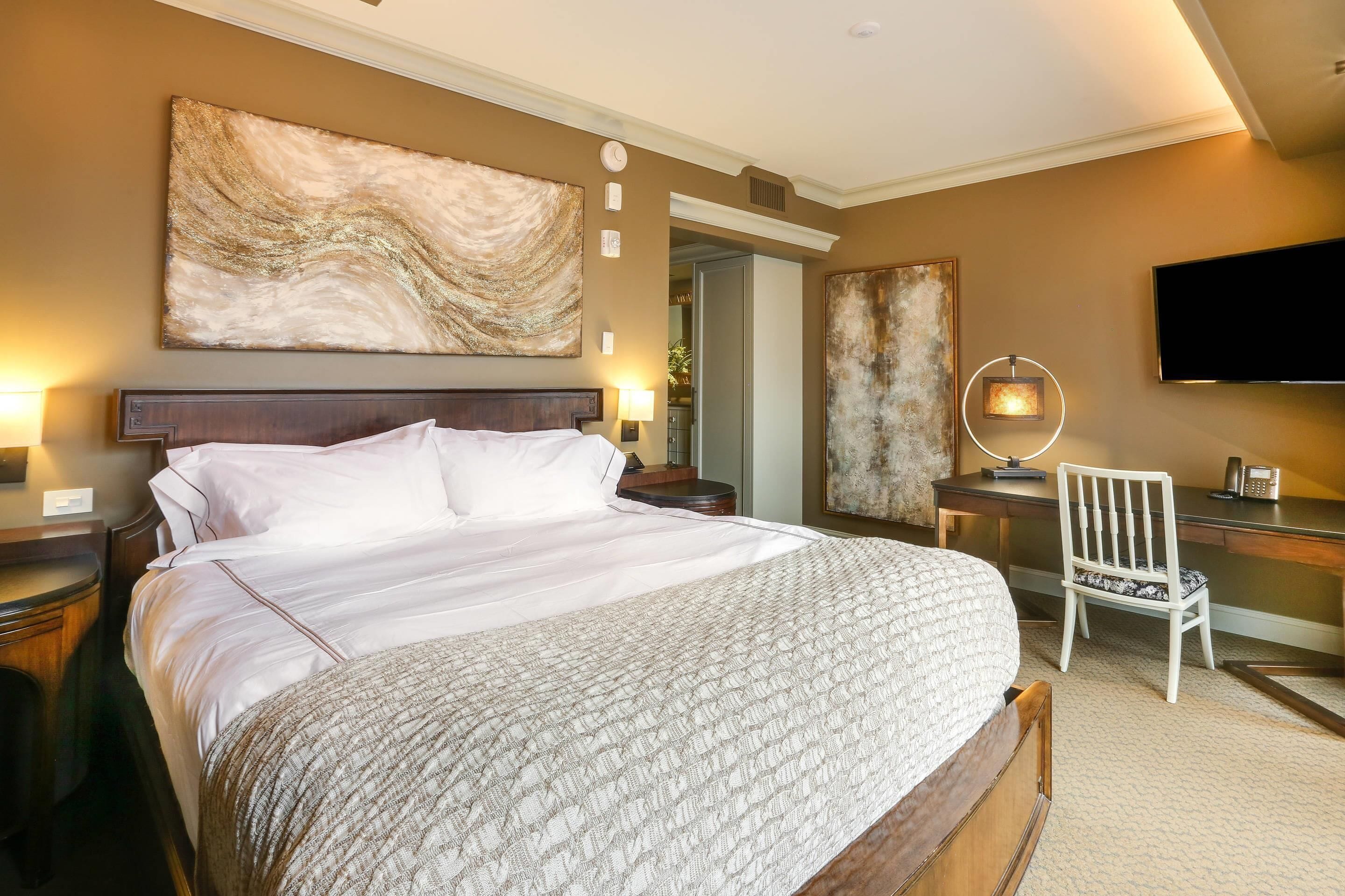 Quarto de hotel com uma cama grande, escrivaninha, cadeira, televisão e obras de arte nas paredes.