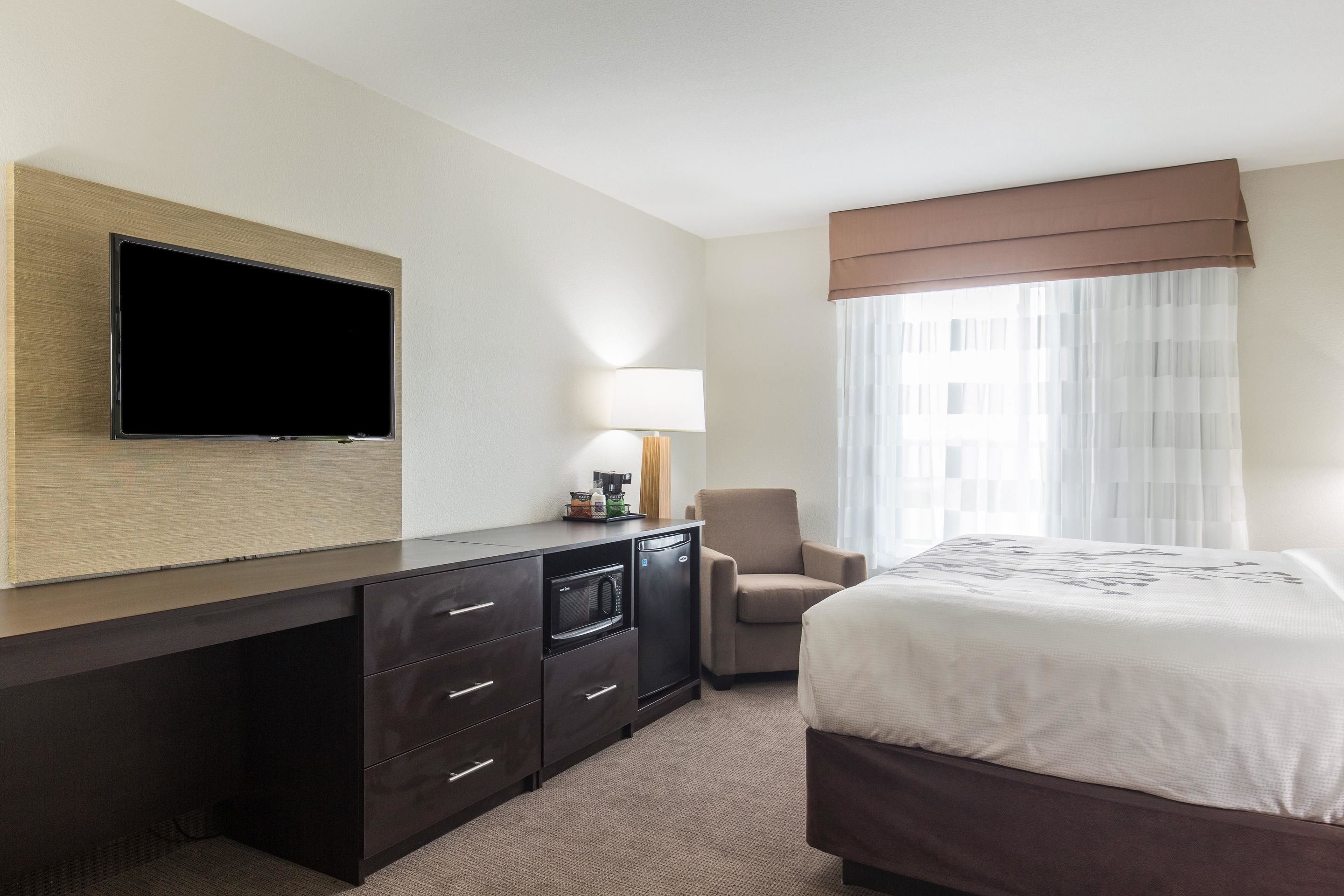 A hotel room with a flat-screen TV mounted on the wall, a desk with a microwave, a chair, and a bed with a large window.