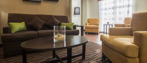 Een moderne hotelkamer met een bruine bank, een ronde salontafel en twee fauteuils. Er hangen afbeeldingen in lijsten aan de muur en er is een groot raam met gordijnen.