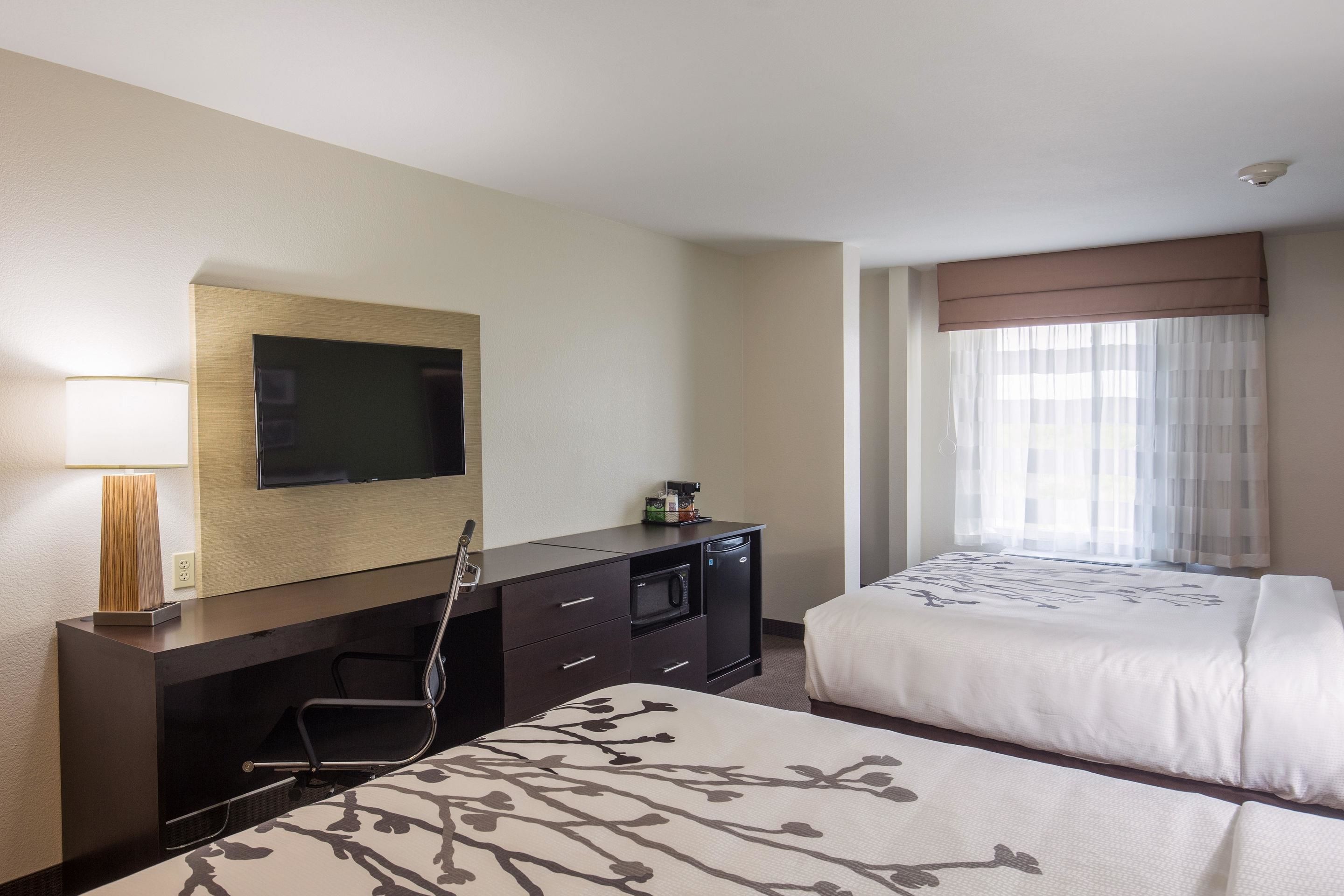 A hotel room with two beds, a TV mounted on the wall, a desk with a lamp, and a window with sheer curtains.