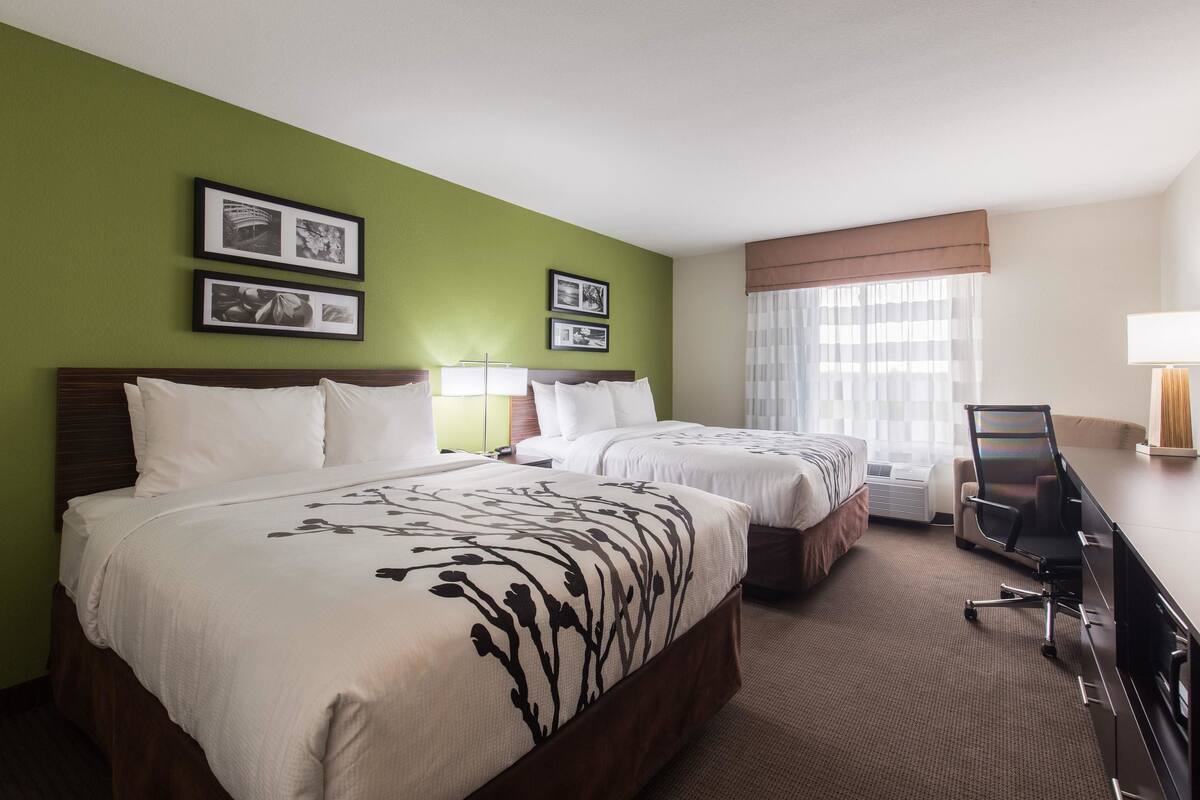A hotel room with two beds, a desk, and a chair. There are framed pictures on the wall and a window with curtains.