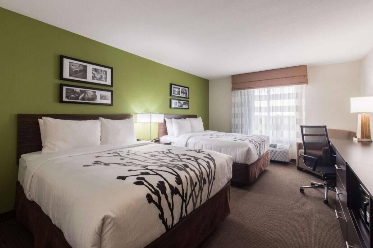 A hotel room with two beds, a desk, a chair, and framed pictures on the wall.