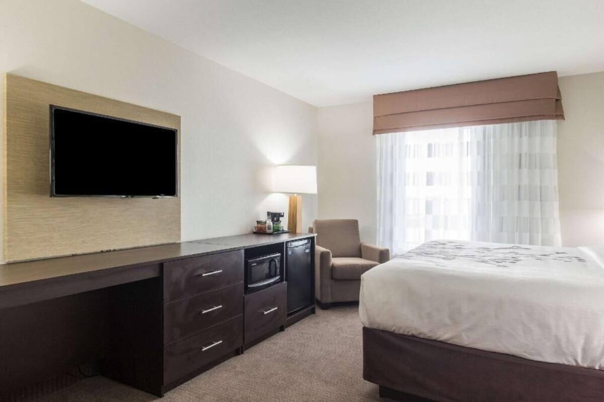 A hotel room with a bed, a desk with a flat-screen TV, a lamp, a chair, and a window with curtains.