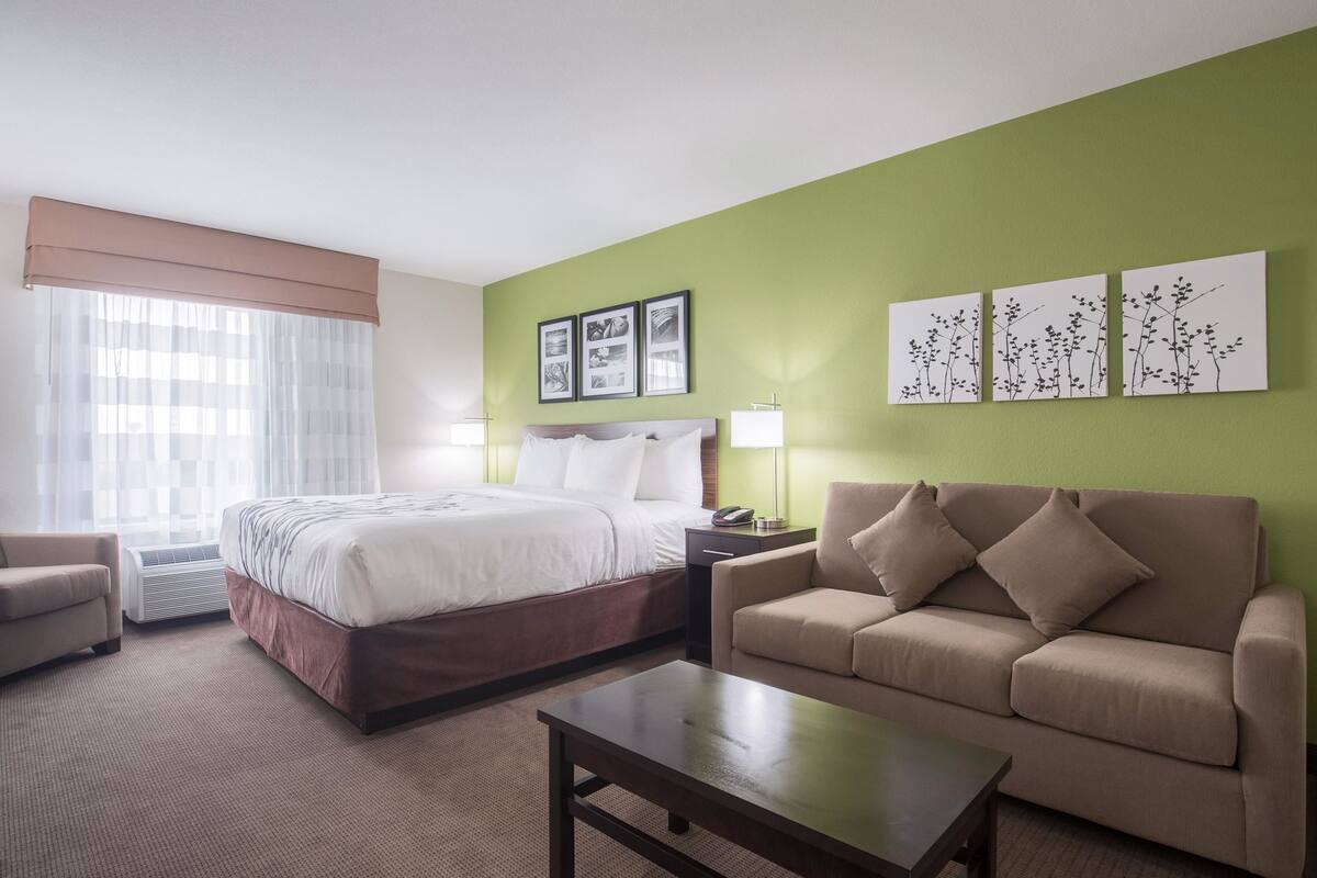 A hotel room with a bed, a sofa, a coffee table, and a desk with a lamp. There are three framed pictures on the wall and a window with sheer curtains.