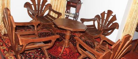 Un comedor con una mesa redonda de madera rodeada por seis sillas de madera. En el centro, se encuentra una pequeña mesa auxiliar de madera con un jarrón de flores. El suelo está cubierto por un tapete estampado.