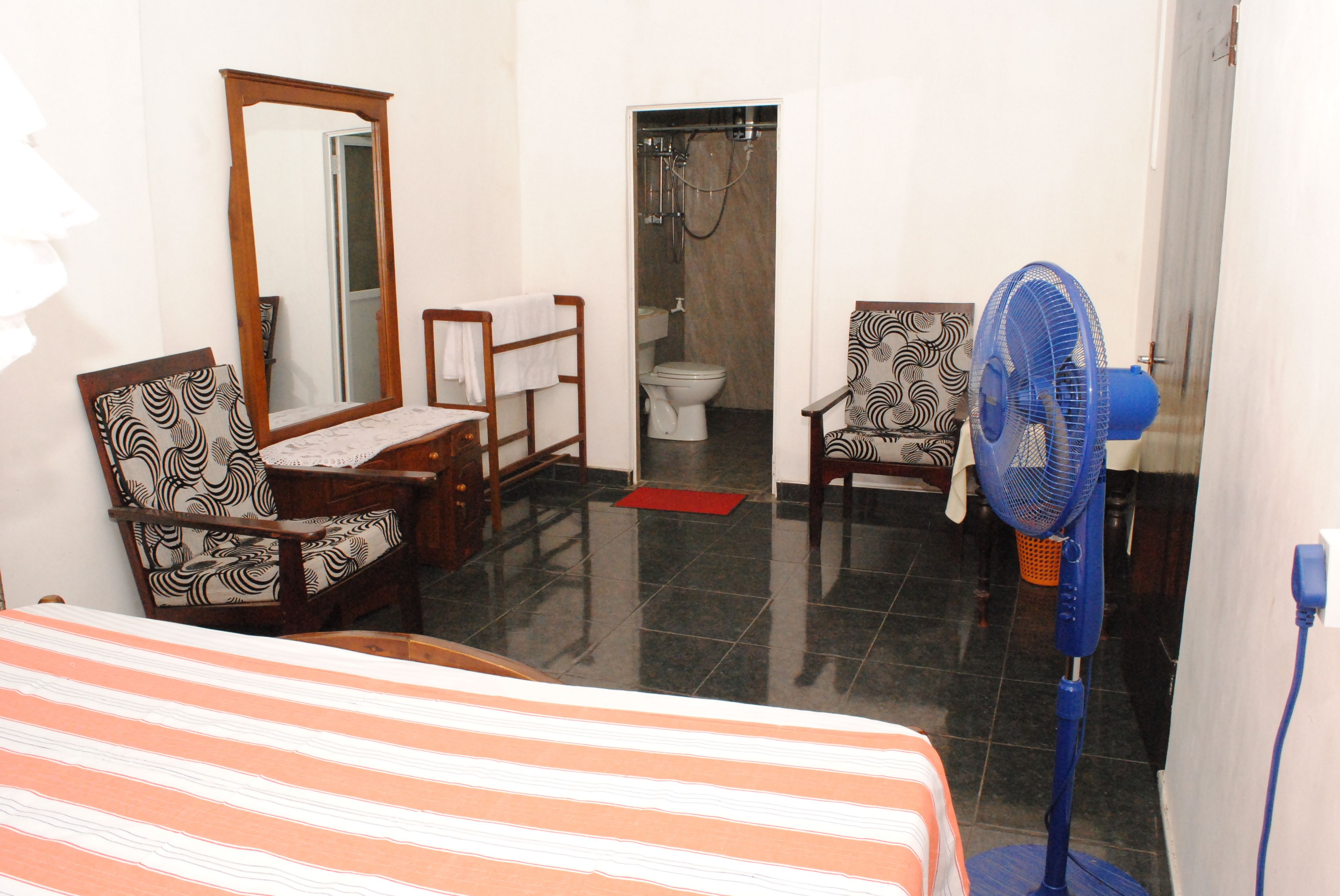 A hotel room with a bed, two chairs, a table, a mirror, a fan, and a bathroom with a shower.