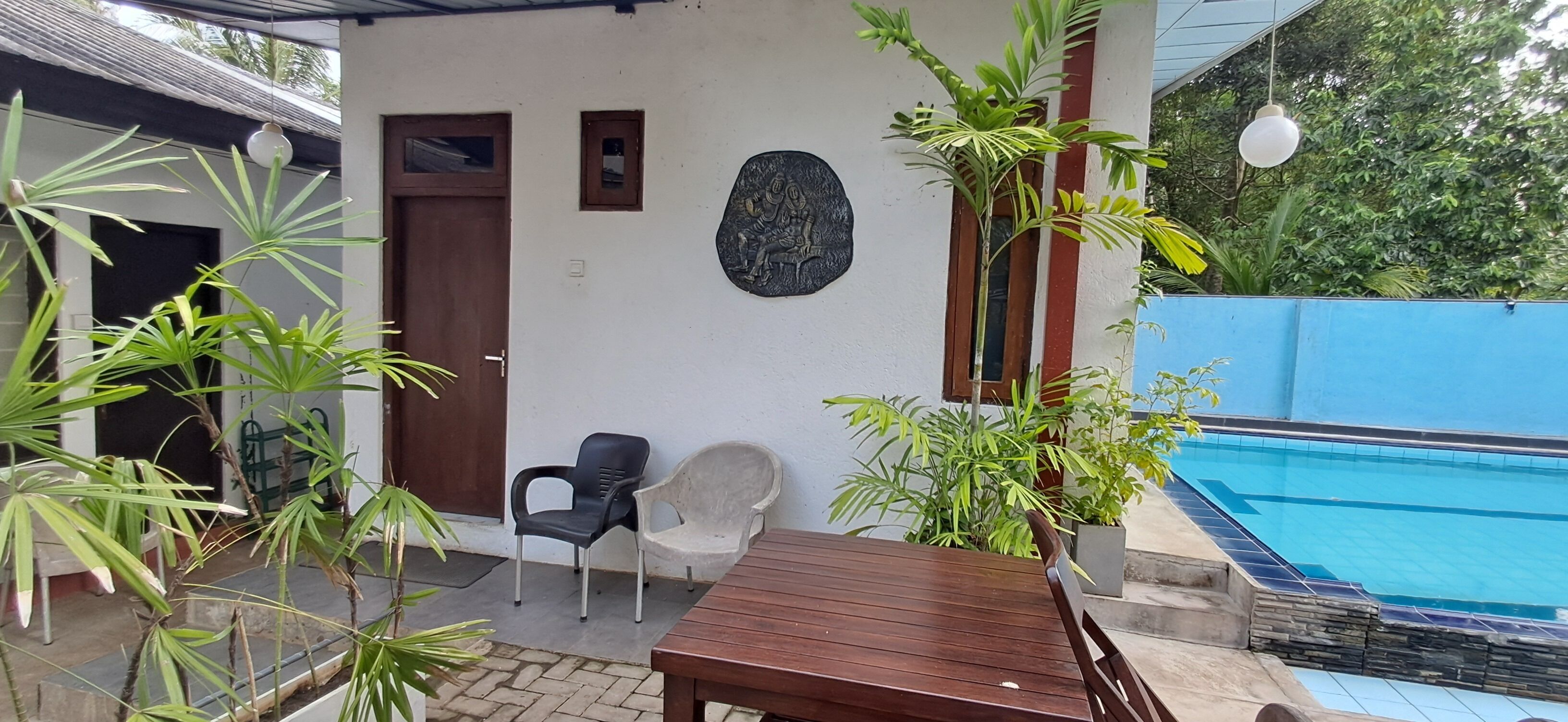 A poolside area with a wooden table and chairs, a small statue on the wall, and a blue fence.
