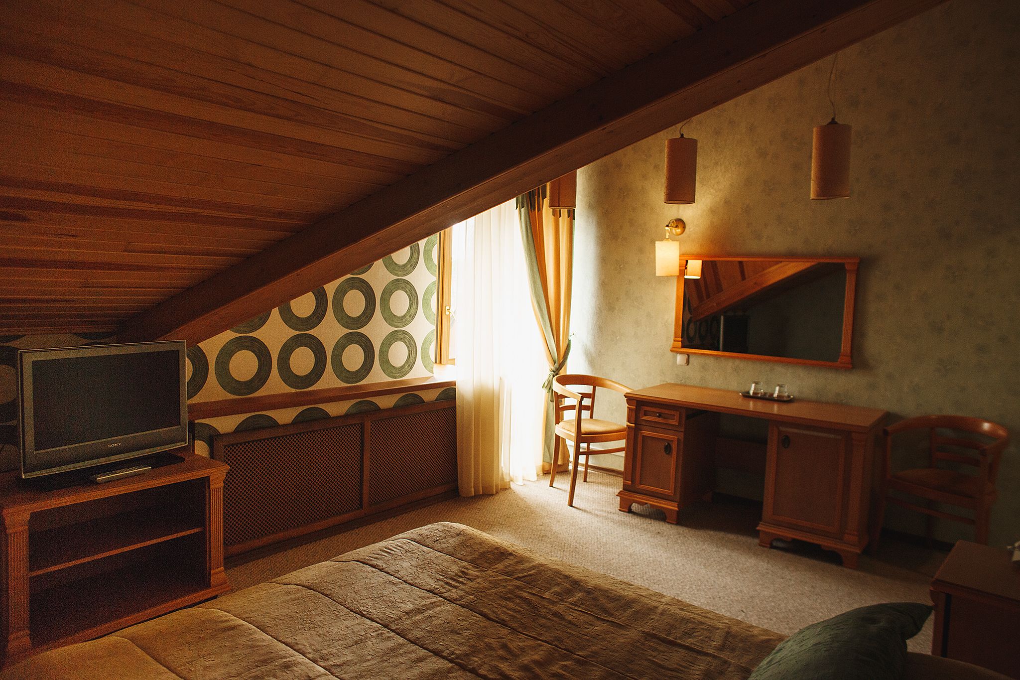 A cozy attic bedroom with a wooden ceiling, a television on a wooden stand, a desk with a mirror, and a patterned wall.