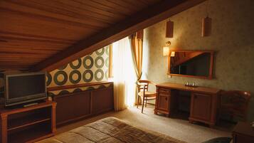 A cozy attic bedroom with a wooden ceiling, a television on a wooden stand, a desk with a mirror, and a patterned wall.