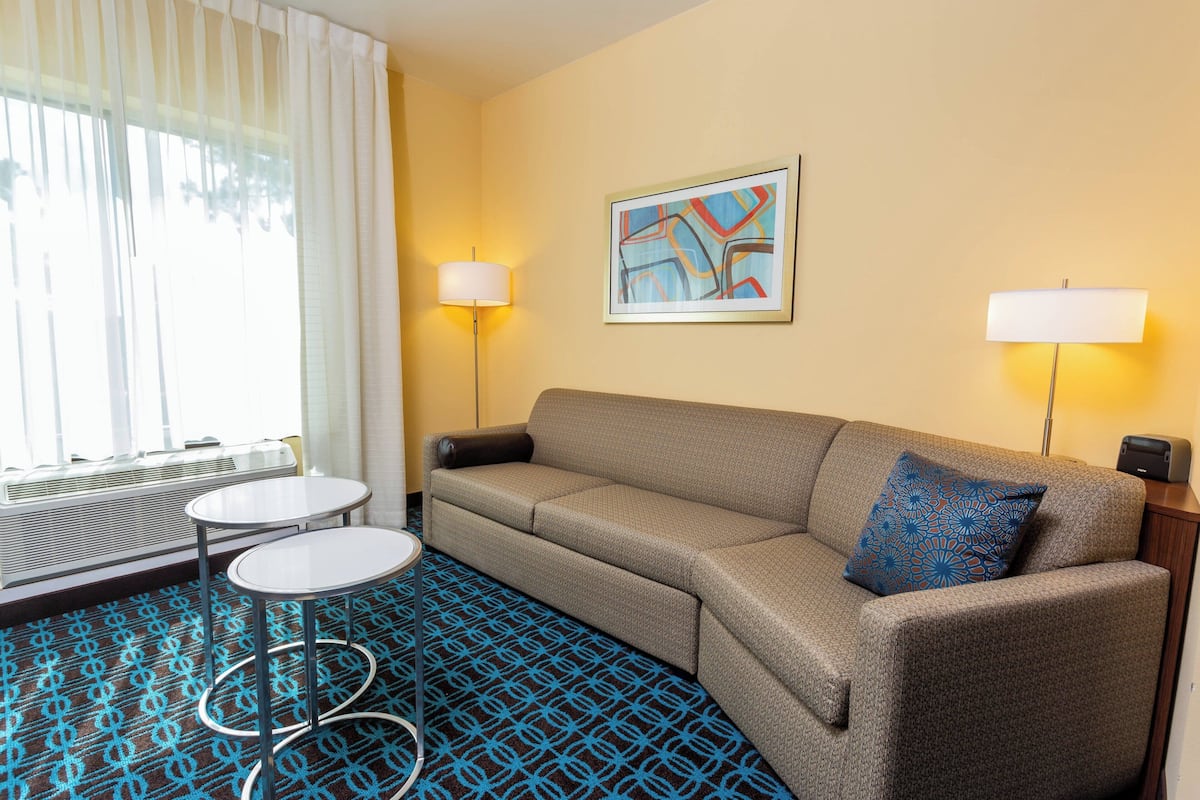 A hotel room with a sofa, two side tables, a patterned carpet, a radiator, and a framed abstract painting on the wall.