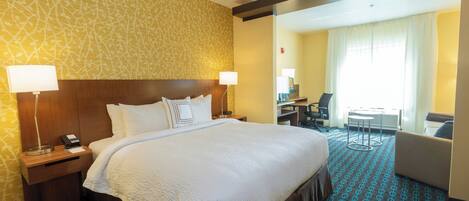 Quarto de hotel com uma cama grande, duas luminárias de cabeceira, uma escrivaninha com cadeira e vista para uma área de refeições através de um painel de vidro.