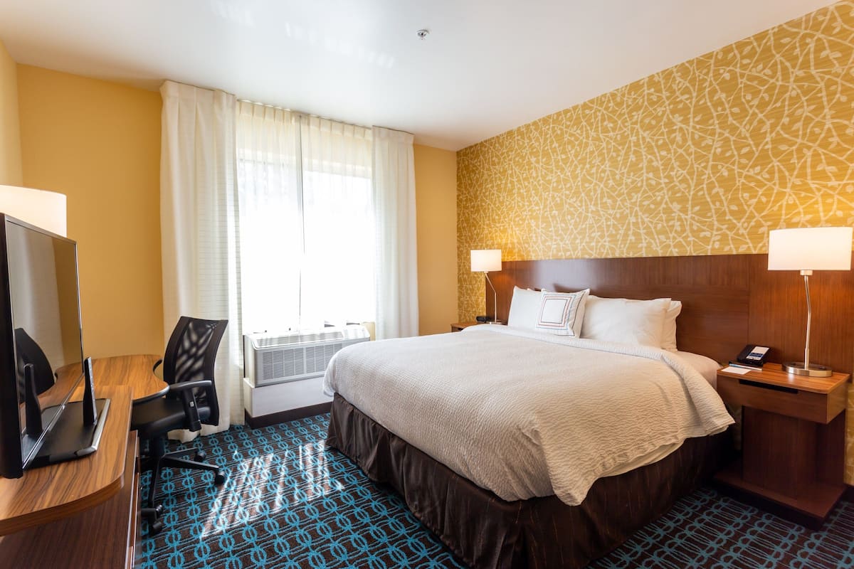 A hotel room with a bed, a desk with a computer, a chair, a window with curtains, and a patterned wall.