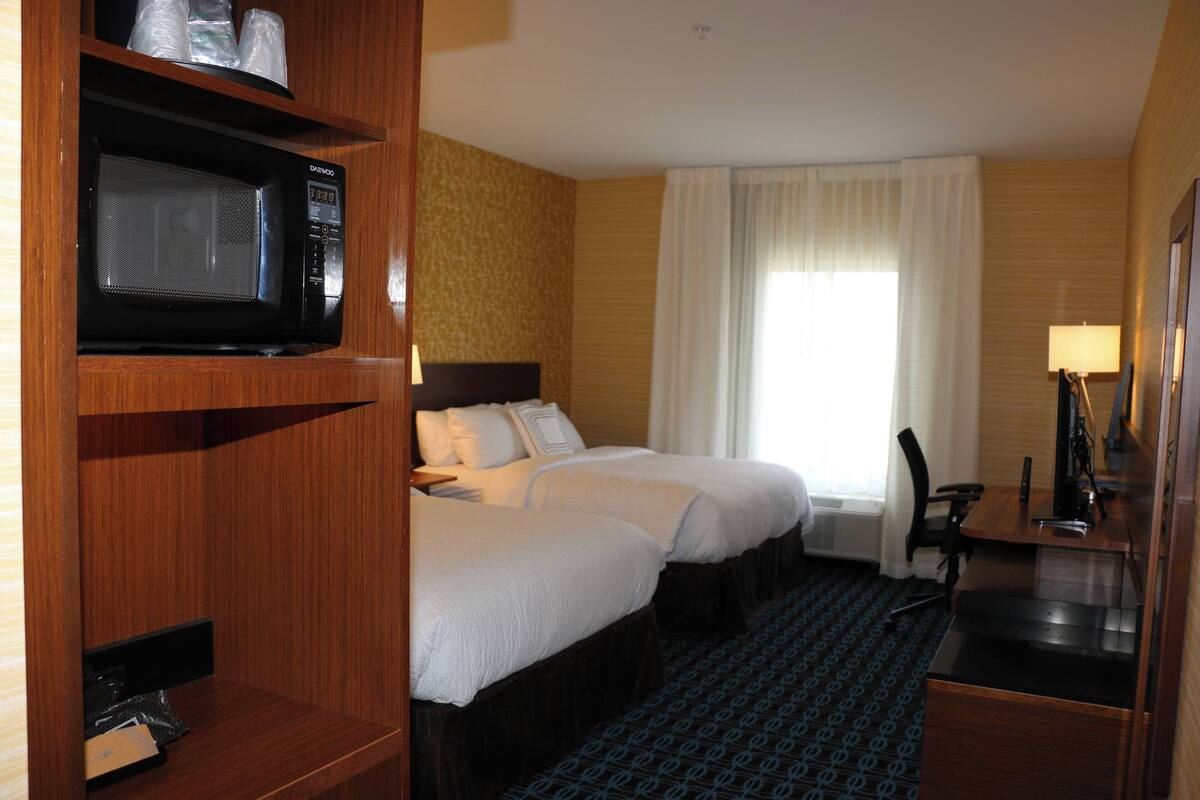 A hotel room with two beds, a desk, a chair, a microwave, and a lamp.