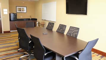 A conference room with a long table, six chairs, a whiteboard, a flat-screen TV, and a kitchenette in the background.