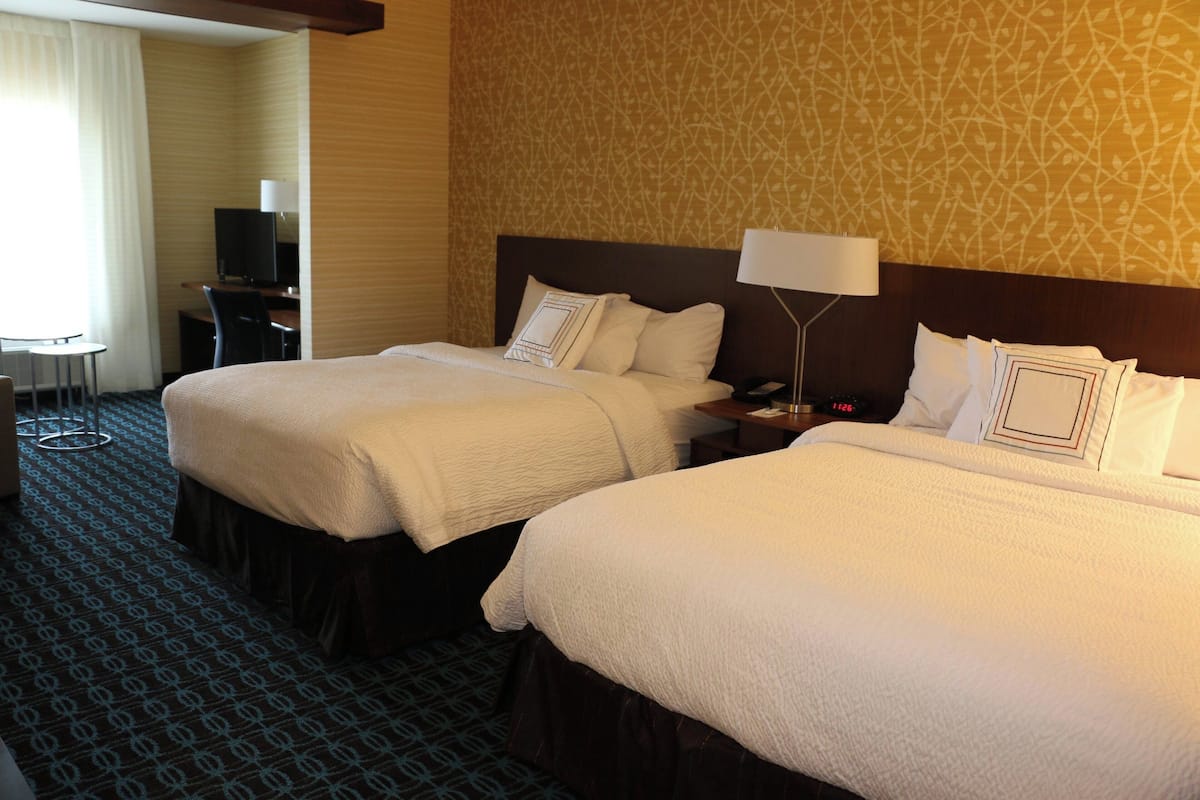 A hotel room with two beds, a desk, a chair, a TV, and a lamp.