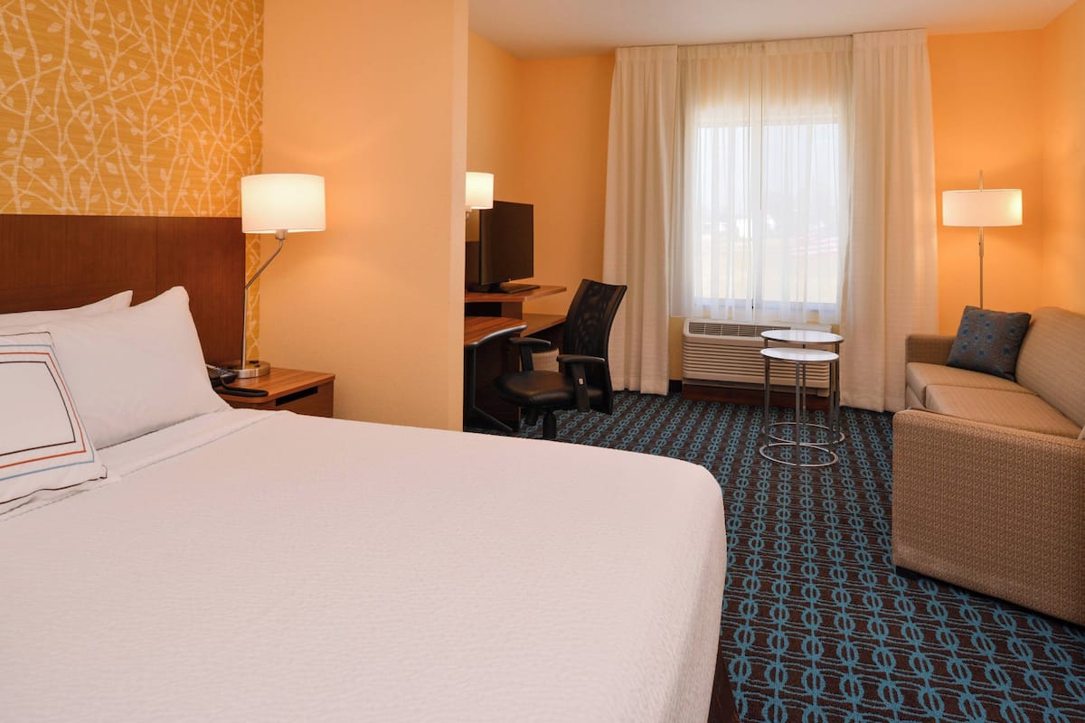 A hotel room with a large bed, a desk with a chair, a small table, a sofa, and a window with curtains.
