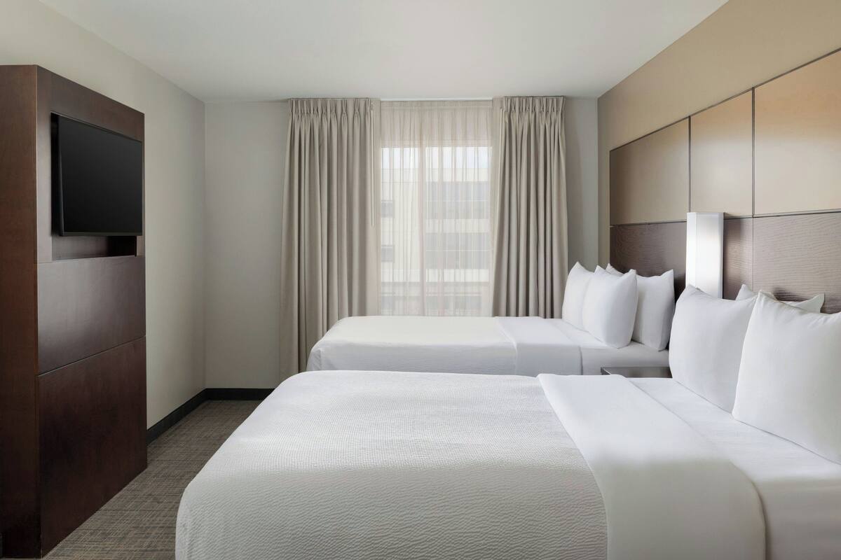 A hotel room with two beds, a flat-screen TV mounted on the wall, and a large window with curtains.