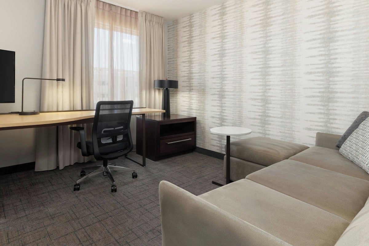 A hotel room with a desk, chair, TV, lamp, sofa, and curtains.