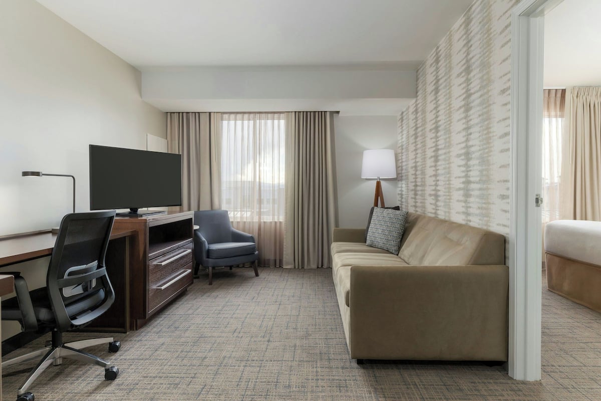 A hotel room with a desk, chair, television, sofa, and curtains.