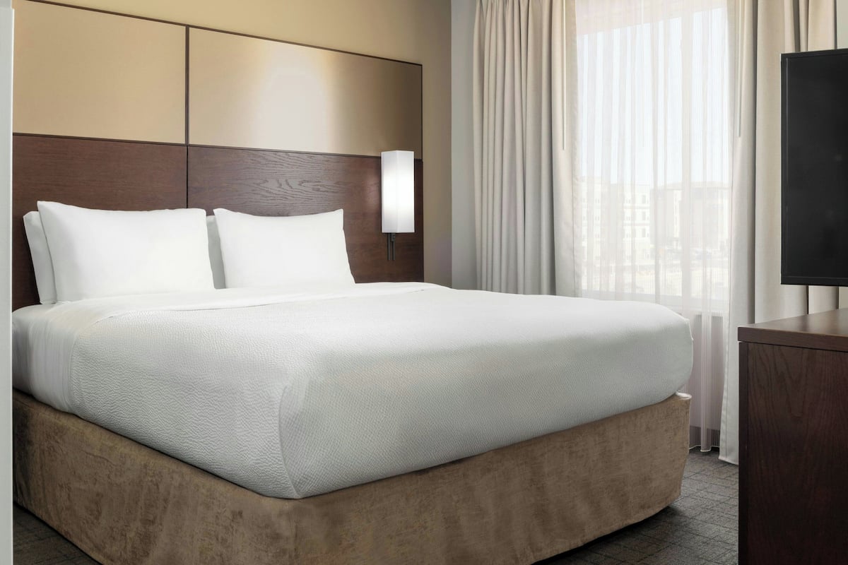 A neatly made bed with a wooden headboard and a bedside lamp, a large window with sheer curtains, and a flat-screen TV on a wooden stand.