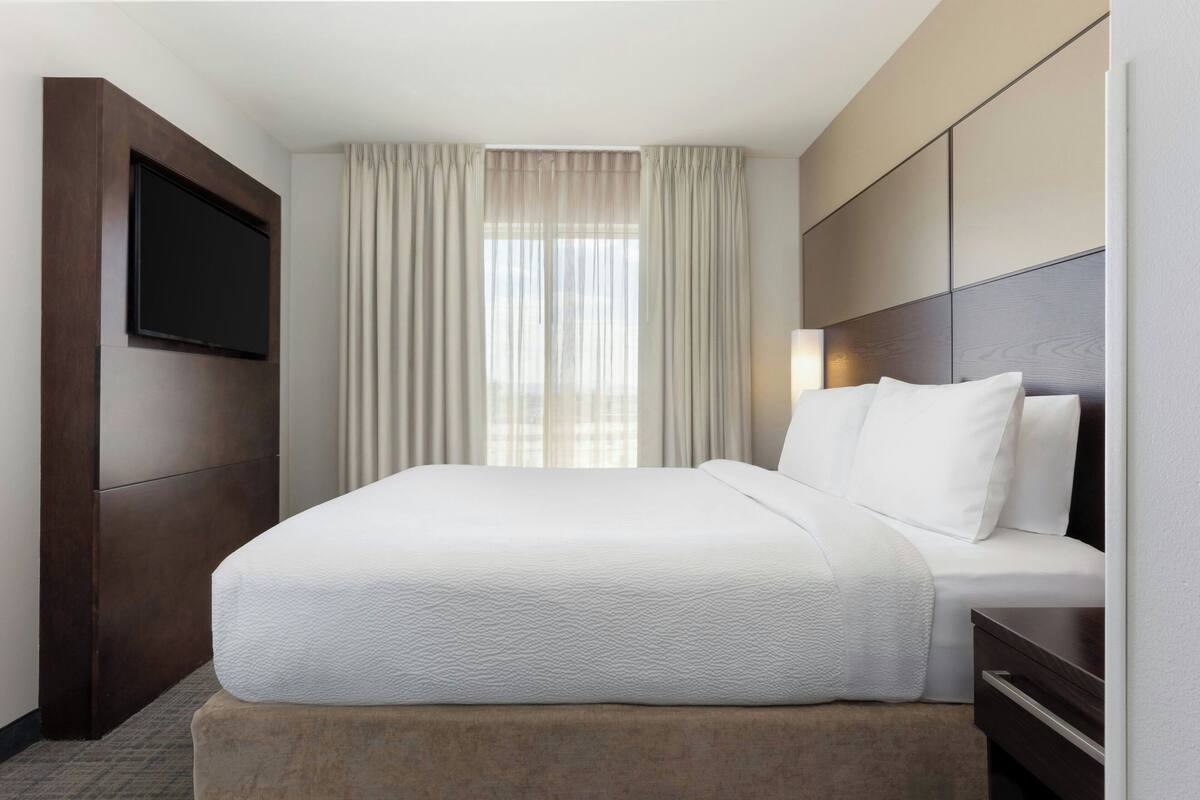 A hotel room with a large bed, a flat-screen TV mounted on the wall, and a window with sheer curtains.