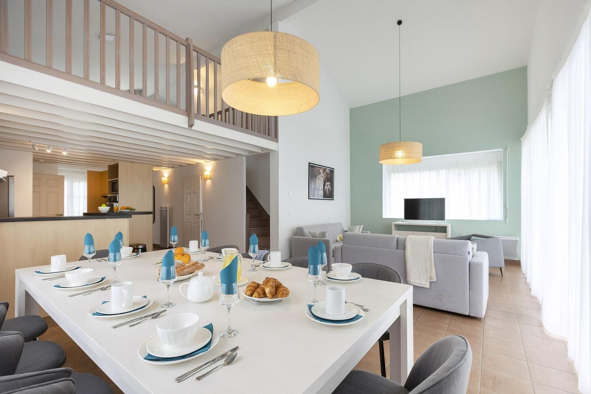 Un espace repas moderne avec une table blanche dressée pour le petit-déjeuner, un escalier menant à un niveau supérieur et un salon avec un canapé gris et un téléviseur.
