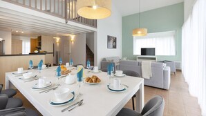 A modern dining area with a white table set for breakfast, a staircase leading to an upper level, and a living room with a grey sofa and a television. - Villa in France with Golf Course & Pool (Gujan Mestras)
