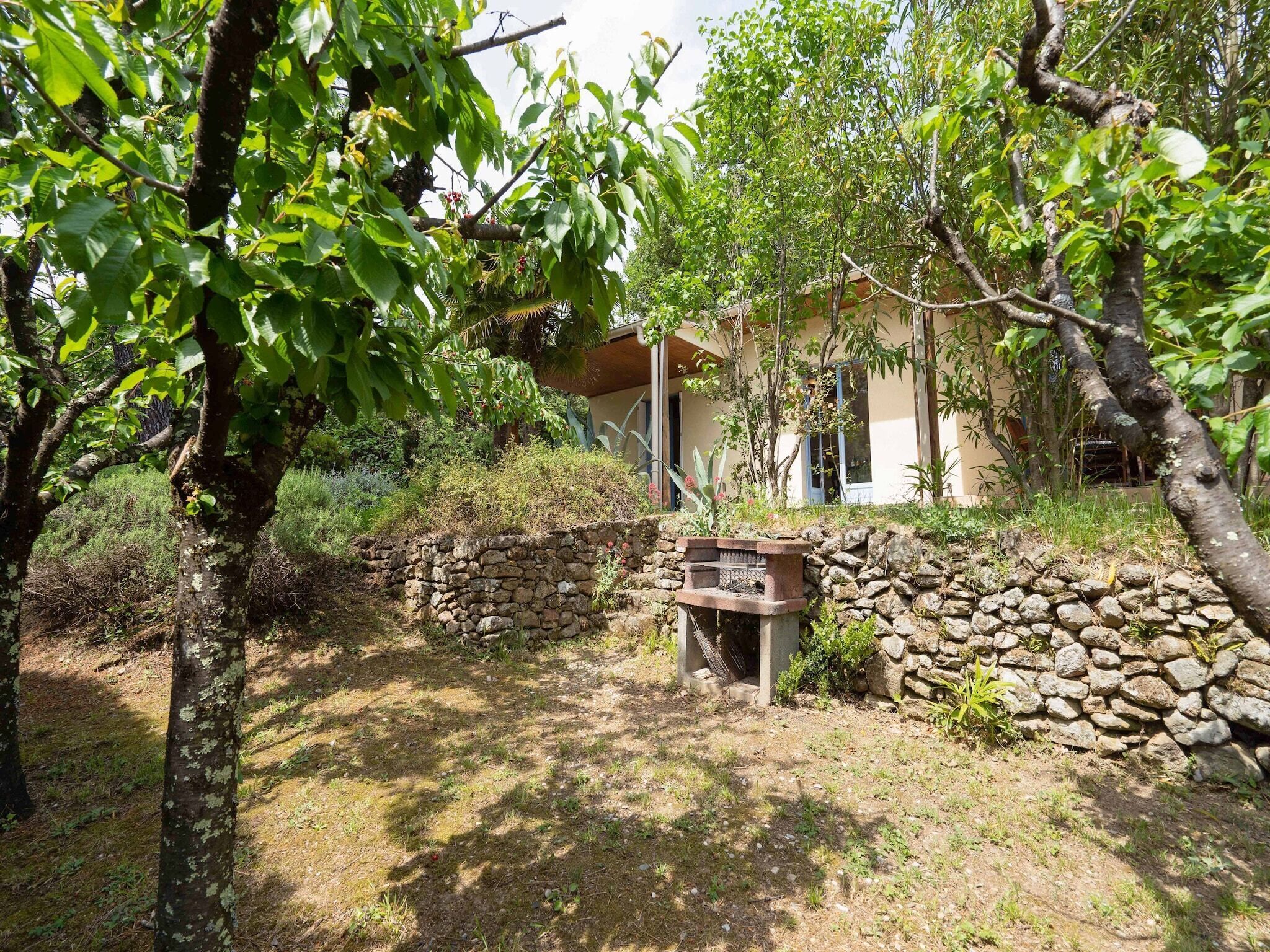 A rustic stone wall with a wooden bench in a garden area, surrounded by trees and a building with a sloped roof.