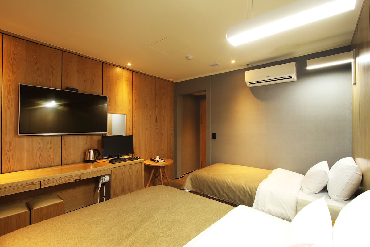 A hotel room with a wooden desk, a flat-screen TV, a bed with white linens, and an air conditioning unit on the wall.