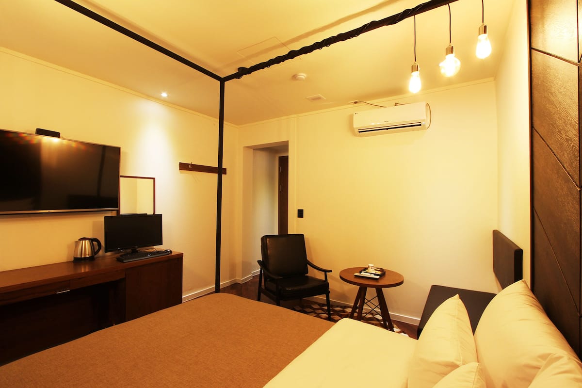 A hotel room with a bed, a desk with a television, a chair, a small table, and a wall-mounted air conditioner.
