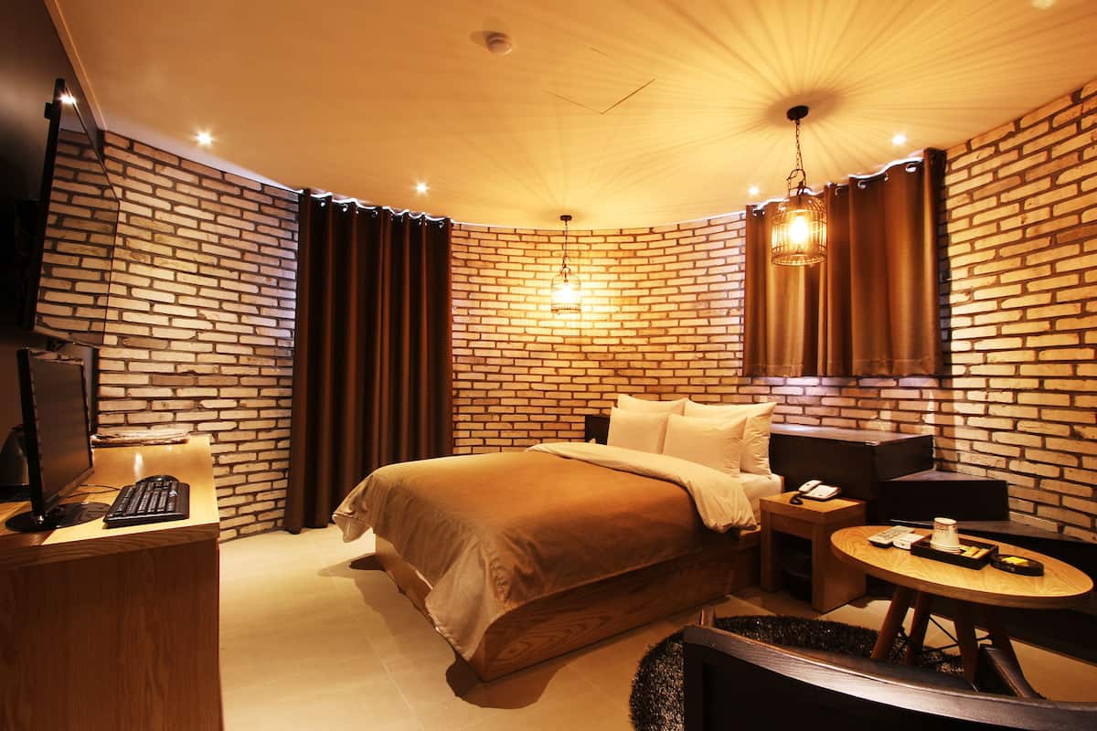 A hotel room with a brick wall, a bed, a desk with a computer, a round table, and a hanging light fixture.