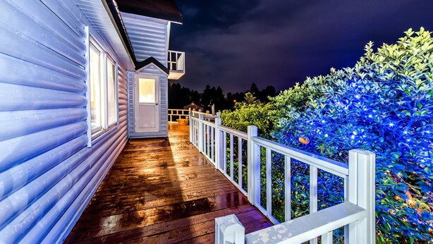 Een houten terras met een witte balustrade, een tuinhuisje en een goed verlichte tuin.