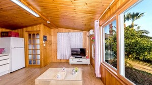 Een compact houten vakantiehuisje met kitchenette, televisie en een groot raam met uitzicht op groen.