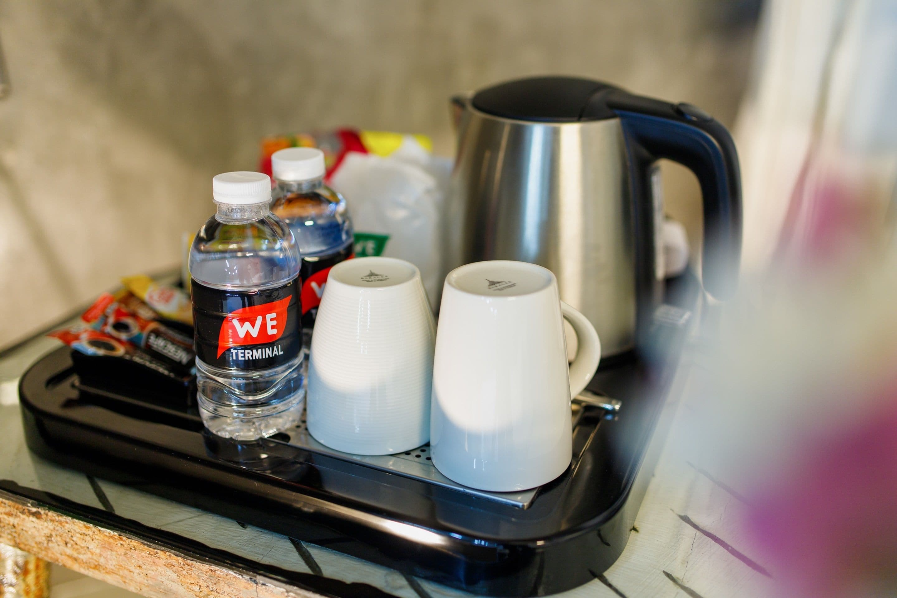 Una bandeja con una botella de agua, dos tazas blancas y un hervidor de acero inoxidable.