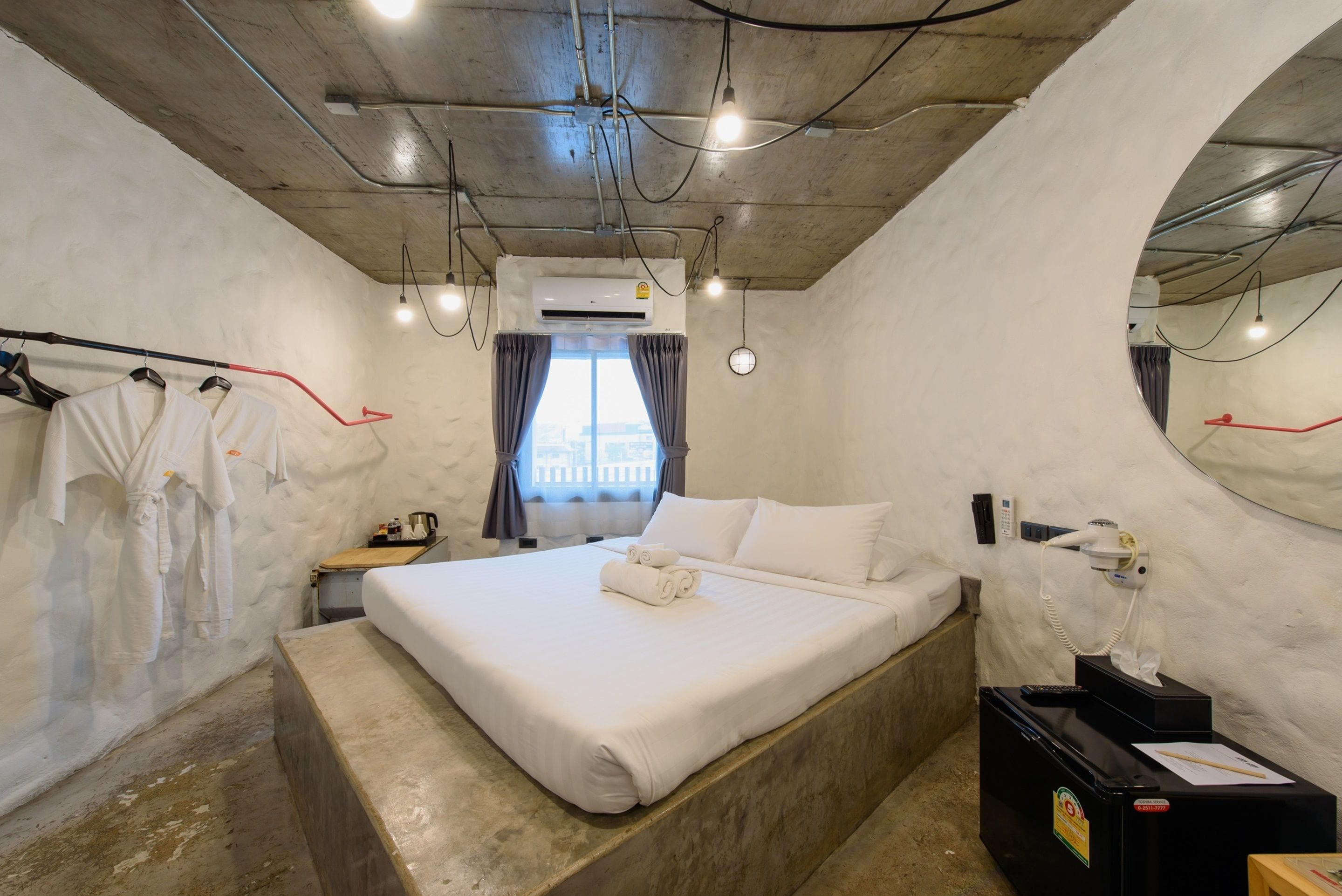 A hotel room with a bed, a bedside table, a mirror, an air conditioning unit, and a window with curtains.