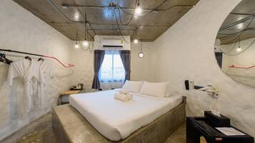 A hotel room with a bed, a bedside table, a mirror, an air conditioning unit, and a window with curtains.
