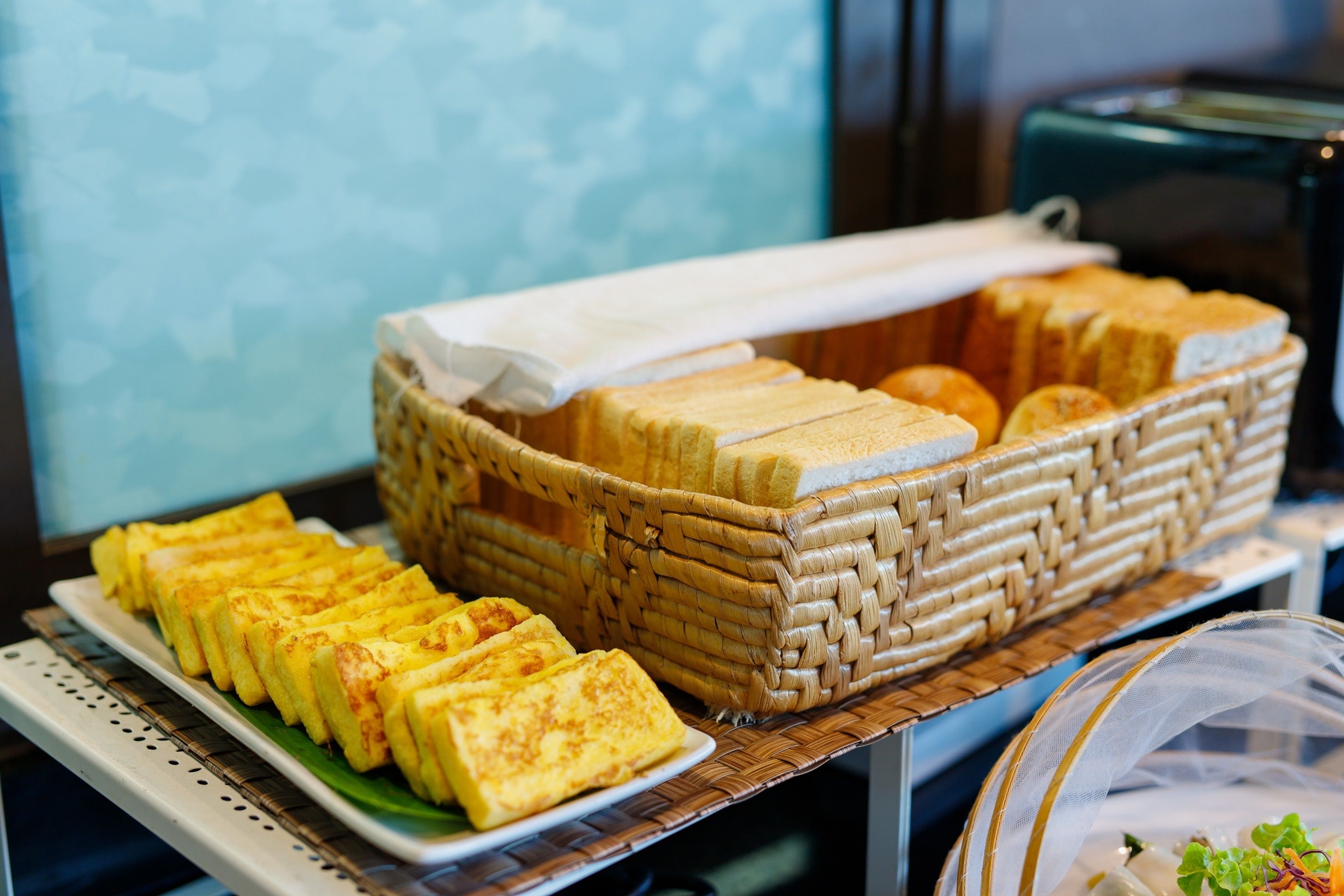 A buffet with a variety of food items including sandwiches and pastries, arranged on a table with a woven basket and a clear plastic cover.