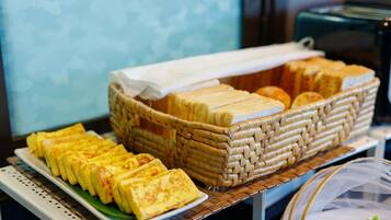 A buffet with a variety of food items including sandwiches and pastries, arranged on a table with a woven basket and a clear plastic cover.