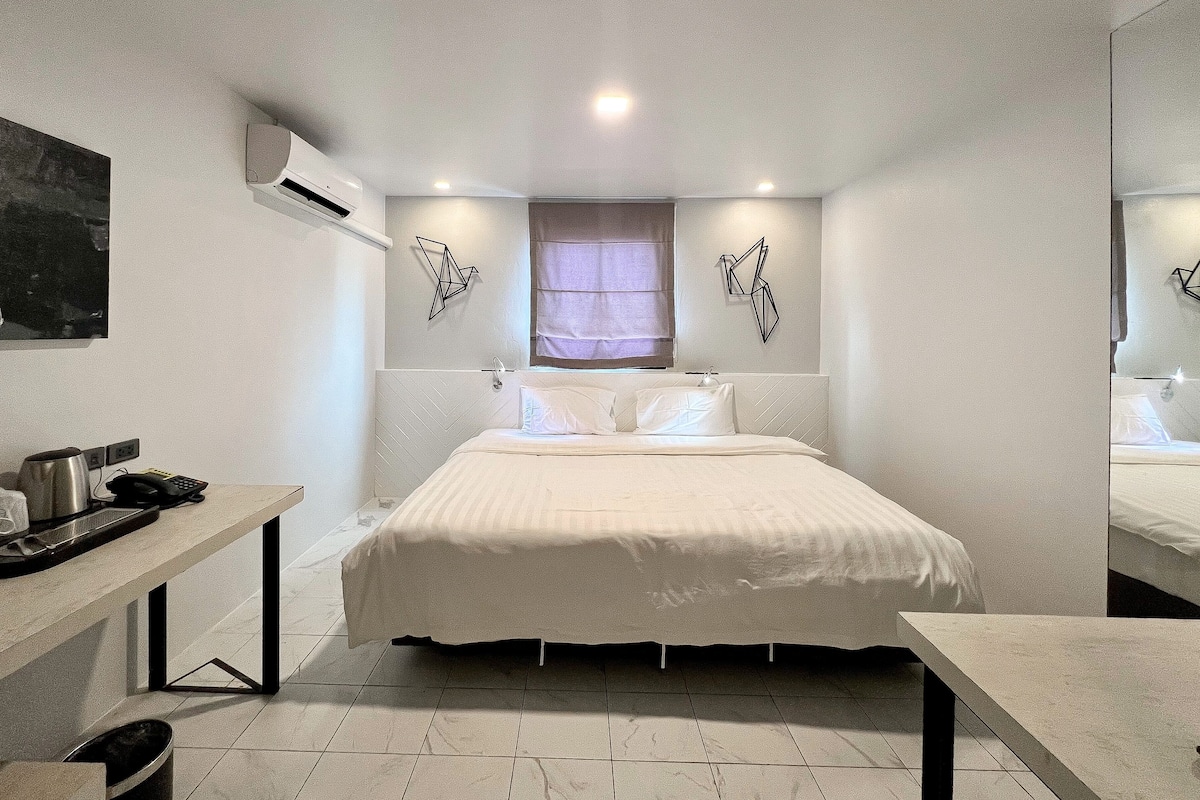 A bedroom with a bed, bedside tables, a desk, an air conditioning unit, and a window with curtains.