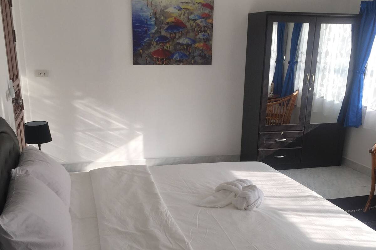 A neatly made bed with a white comforter and a folded towel, a dark-colored wardrobe, a painting on the wall, and a window with blue curtains.