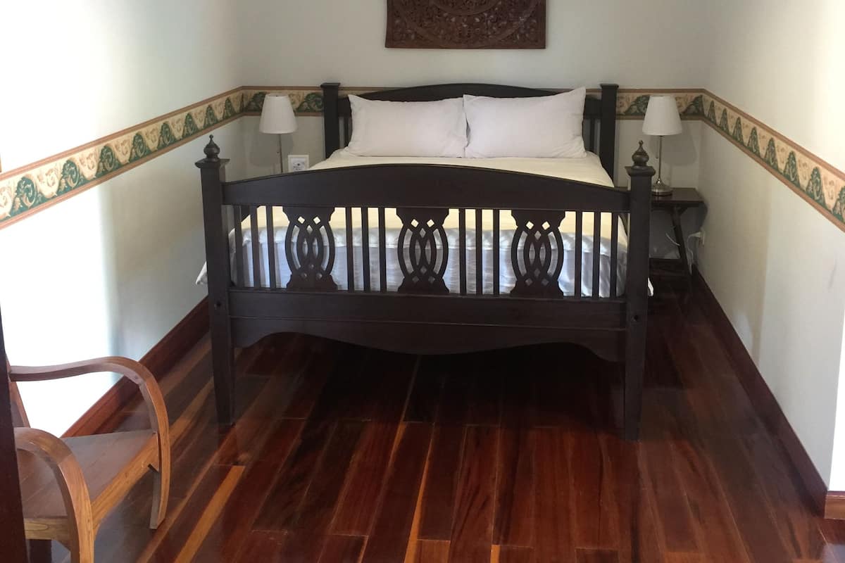 A bedroom with a wooden bed, white bedding, a decorative wall hanging, and a wooden chair.