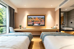 Ein Hotelzimmer mit zwei Betten, einem Schreibtisch mit Computer, einem großen Gemälde eines traditionellen Gebäudes, einem Fenster mit Vorhängen und einem Badezimmer mit Spiegel und Waschbecken.