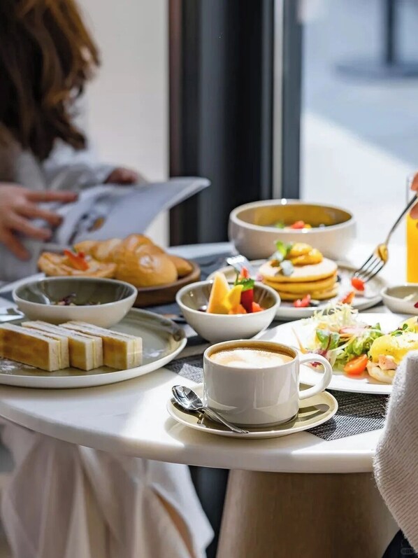 Uma mesa de café da manhã com vários pratos, incluindo panquecas, uma tigela de frutas e uma xícara de café.