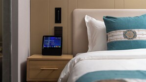 Una cama bien hecha con un almohá de color aguamarina, una mesita de noche con un reloj digital y un panel de pared.
