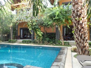 A swimming pool with a surrounding garden, a palm tree, and a building with outdoor seating.